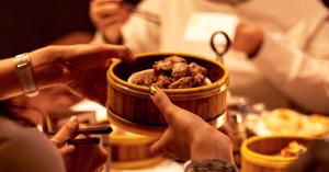 Guests pass a bamboo serving bowl of food around a table during a warmly lit family-style dinner.