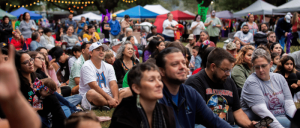A diverse group of people seated outdoors at a community event with tents and string lights in the background.