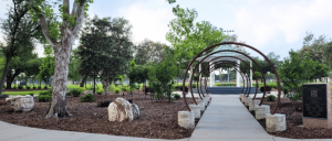 Park walkway with curved metal arches, trees, rocks, and a plaque on the right side under a clear sky.