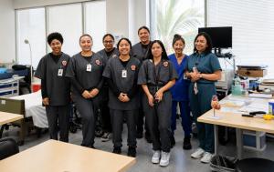 Group photo of students and instructors in scrubs standing together in a medical training classroom with exam beds, desks, and clinical equipment.