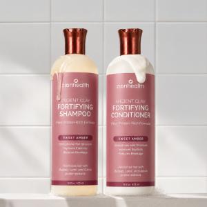 Zion Health Fortifying Shampoo and Conditioner standing side by side on a white bathroom counter against glossy white tiles, with shampoo and conditioner textures dripping over the bottles. The duo features a revolutionary vegan-like keratin system design