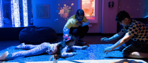 Children play on a projected interactive floor while two adults supervise in a dimly lit, colorful museum exhibit.