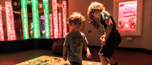 Two children explore an interactive light exhibit in a dimly lit children’s museum.