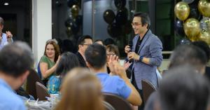 James C Foo Leong speaks into a handheld microphone and gestures while engaging an audience at a professional event, with yellow and black balloons in the background.