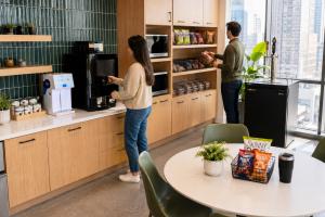 Two employees using a modern office breakroom, one drawing coffee from a bean-to-cup brewer and the other selecting snacks from stocked shelves, with a water dispenser, tap unit, and city view alongside