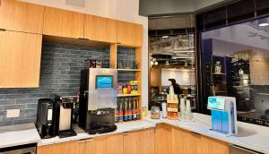 Two employees using a modern office breakroom, one drawing coffee from a bean-to-cup brewer and the other selecting snacks from stocked shelves, with a water dispenser, tap unit, and city view alongside