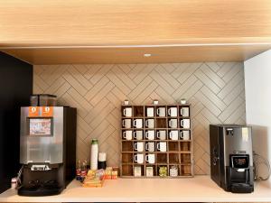 Office breakroom coffee station with two commercial bean-to-cup brewers flanking a wooden mug rack, with cups, syrups, tea, and sweeteners arranged on the counter beneath a herringbone tile wall