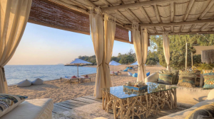 Beachfront lounge with shaded seating, curtains, and ocean views at sunset.