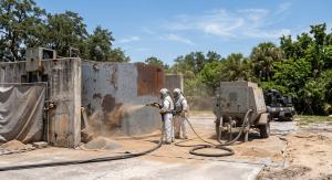 A worker in a white suit applies sand to a concrete wall using a sandblasting technique for surface finishing.