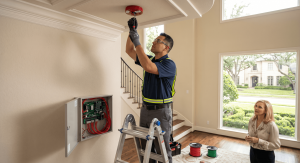 A female technician in a blue polo with high-visibility stripes and safety glasses stands on a metal ladder, using tools to work inside an open gray electrical control panel with many red wires mounted on a beige wall.