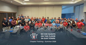 Group photo of dozens of Sleep in Heavenly Peace chapter volunteers and leaders standing and seated around conference tables in a training room, smiling for a group photo. Text overlay reads "SHP, Sleep in Heavenly Peace: No Kid Sleeps On The Floor In Our