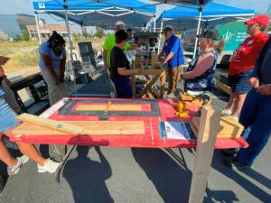 Volunteers gather around an outdoor workstation under a blue canopy tent, assembling wooden bed frame pieces on a red table labeled "Tap Station." A drill press and lumber are set up nearby, with an SHP-branded box truck reading "No Kid Sleeps On The Floo