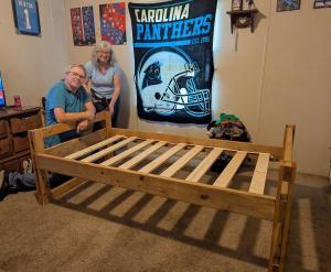 A man and woman smile while kneeling and standing behind a newly assembled wooden bed frame in a bedroom, part of a bed delivery experience during SHP chapter training. A Carolina Panthers banner and sports posters decorate the wall behind them.
