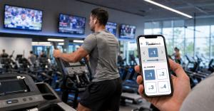 Gym member uses cardio equipment beneath wall-mounted televisions while the AudioFetch app displays selectable TV audio channels on a smartphone.