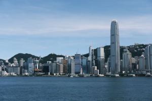 Hong Kong city skyline over Victoria Harbour on a clear day, with numerous skyscrapers and green hills in the distance.