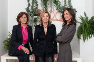 Bacana Communications founders Maria, Irene and Cristina Aguilar pose together against a backdrop of hanging greenery and a neon sign at the agency's office. Three women, the founders of Bacana Communications, stand and sit together smiling for a group p