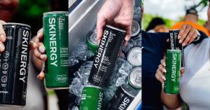 Two hands holding cans of Skinergy zero-sugar energy drink, one black Wild Berry can and one green Green Apple flavor can, each labeled with collagen, caffeine and vitamins.