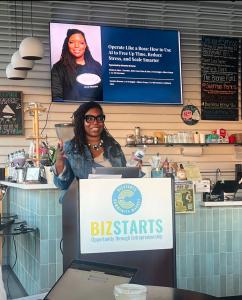 Natasha Broxton speaking at a BizStarts podium while a presentation about using AI to operate and scale a business appears behind her.