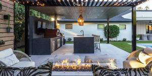 Covered outdoor kitchen with a stone island, built-in grill and pizza oven beside a lit fire pit and lounge seating at a Phoenix Arizona home at dusk