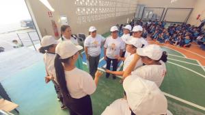Plushie Planet volunteers in white caps with students seated across a school sports court