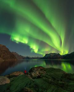 Aurora borealis glows bright green above a calm mountain fjord, reflecting across the water as a lone person in a red jacket stands on the rocky shoreline beneath the night sky.