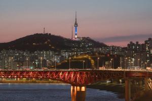 A nighttime photo of the Seoul cityscape. A large, illuminated red bridge crosses the Han River. N Seoul Tower is visible atop a hill in the background, lit in blue and purple, surrounded by numerous apartment buildings under a twilight sky.