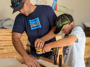 An adult male volunteer in a blue Bunks Across America t-shirt helps a young boy wearing safety glasses operate a power drill to assemble a wooden bed frame at an SHP build event.