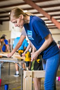 A woman in a blue Bunks Across America t-shirt uses a DeWalt power drill to assemble a wooden bed frame at an indoor SHP build event, with other volunteers working in the background.