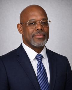Michael D. Walker, PMP®, Cheetah Learning Distinguished Cheetah Alum Award for Significant Impact, sits for a professional picture wearing gold rimmed glasses and a navy suit.