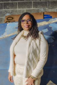 Natasha Broxton standing inside an automotive recycling warehouse with vehicle body panels and parts inventory behind her.