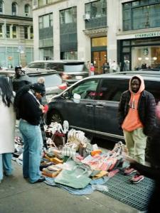 A market vendor handing a wrapped package to a customer across a table of handmade goods at an outdoor New York market.