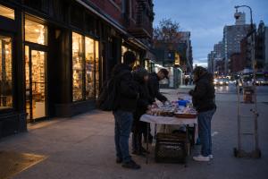 A New York City block at dusk showing lit permanent storefronts alongside a small temporary vendor table on the sidewalk.