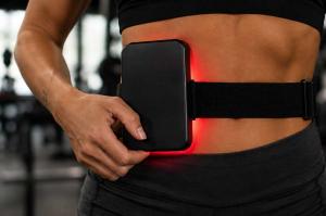 A woman standing in a gym wearing a red light therapy device on her abdomen.