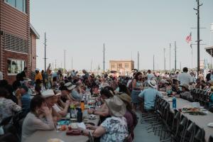 A long outdoor communal dining table filled with seated guests, many wearing cowboy hats, eating and talking under a hazy evening sky. The table stretches into the distance alongside a row of string lights and utility poles, with a brick building on the l