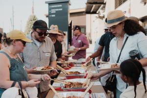 Guests at an outdoor buffet line spoon barbecue chicken and side dishes onto compostable plates from foil catering trays set along a long table, with more guests waiting in line behind them.