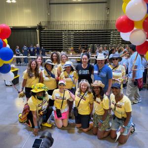 A group of twelve people pose for a photo indoors, most wearing matching yellow "Our Table is a Mile Long" t-shirts and several wearing sun hats, standing and kneeling in two rows beneath balloon arches in red, blue, yellow, and white. Long prep tables an