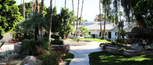 Courtyard with a central fountain, surrounded by palm trees, manicured bushes, green lawn, and a white building in the background.