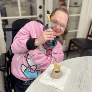 Woman in pink sweatshirt smiling and holding up a paintbrush while sitting indoors near a black chair.