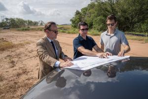 Three men in sunglasses review a large site plan spread across the hood of a car on cleared land at the Occam Foundry campus site in Austin, Texas, with earthmoving equipment working in the background.