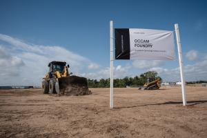 A wheel loader moves dirt on cleared land under a blue sky at the Occam Foundry site in Austin, beside a banner on white posts reading "Future home, Occam Foundry, a quantum technology campus in Austin," with a track loader working behind it.