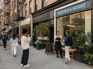 A creator photographing a neighborhood commercial street while a café owner talks with a customer outside her shop.