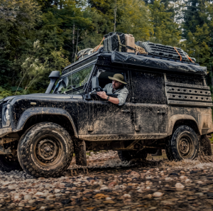 Explorer driving a mud-covered off-road vehicle through rugged forest terrain, wearing a Luminox Adventure Watch on his wrist.