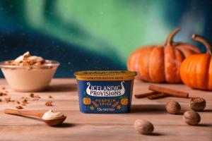 A cup of Icelandic Provisions Pumpkin Spice Skyr with 15g protein on a wooden surface with autumn spices, pumpkins, and a Northern Lights sky in the background