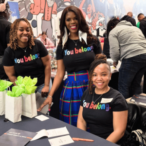 Three smiling women wearing "You belong" shirts stand and sit behind a table with gift bags and papers at a vibrant event.