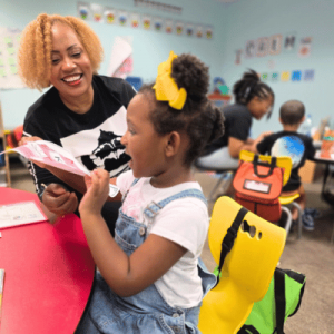 Teacher smiling and reading a book with a young girl wearing a yellow bow in a colorful classroom.