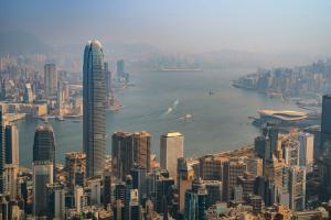 Aerial view of the Hong Kong skyline and Victoria Harbour, featuring the International Finance Centre (IFC) and dense corporate skyscrapers against a hazy mountain backdrop.