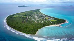 Aerial view of a tropical island with dense green vegetation, a coastal village, and an enclosed harbor, surrounded by bright blue ocean water and coral reefs.