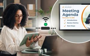 Woman using a tablet and laptop beside an Amazon Signage Stick connected to a display showing a meeting agenda, illustrating digital signage management and connectivity.