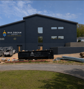 Exterior view of the BHA Dream Center under construction in Bessemer, Alabama. The dark blue building features the BHA Dream Center logo, with construction equipment, materials and a dumpster visible in front as work continues on the facility.