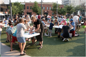 Children and adults gather around tables to play games at a busy outdoor event in a downtown plaza.